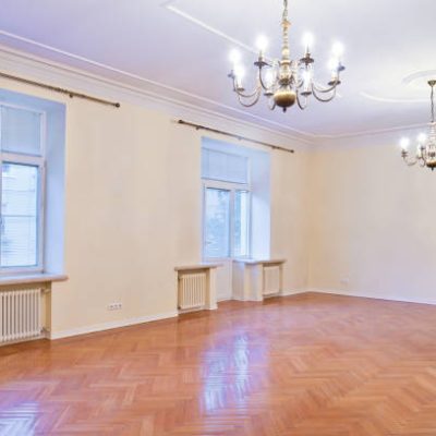 A huge empty room without furniture with laminate flooring high ceilings and chandeliers.